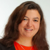 Frau Tratzmüller mit schulterlangem, braunem, gewelltem Haar, trägt ein rotes Oberteil und eine silberne Halskette